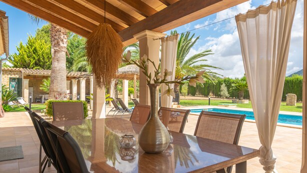 An outdoor dining area with a wooden table, chairs, a vase, and a view of a swimming pool and greenery.