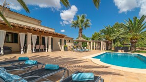A poolside area with sun loungers, a swimming pool, palm trees, and a building with a tiled roof.