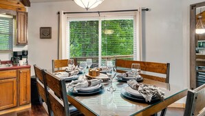 Zona pranzo con tavolo in vetro apparecchiata per quattro, sedie in legno e vista sul verde attraverso le persiane.