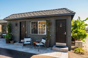 A small, single-story building with a dark gray exterior and a tiled roof. It features two doors, one with a window, and a small porch area with a bench and potted plants.