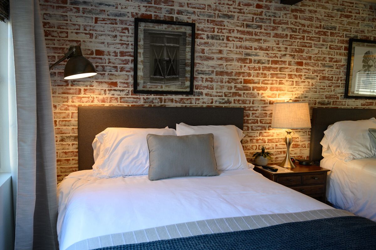 A hotel room with a brick wall, a large bed with white linens, two bedside tables with lamps, and framed pictures on the wall.