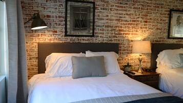A hotel room with a brick wall, a large bed with white linens, two bedside tables with lamps, and framed pictures on the wall.
