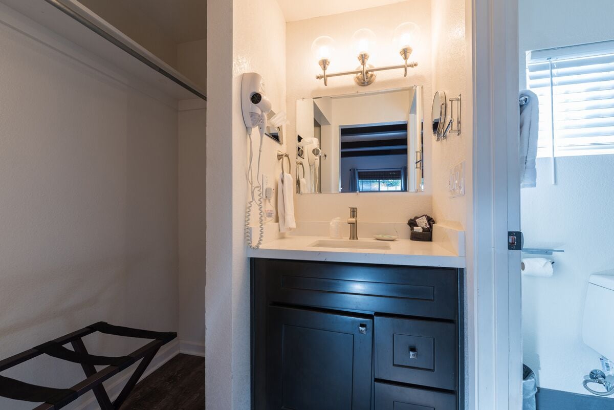 A bathroom with a white sink, dark cabinets, a mirror with lights, and a hairdryer on the wall.