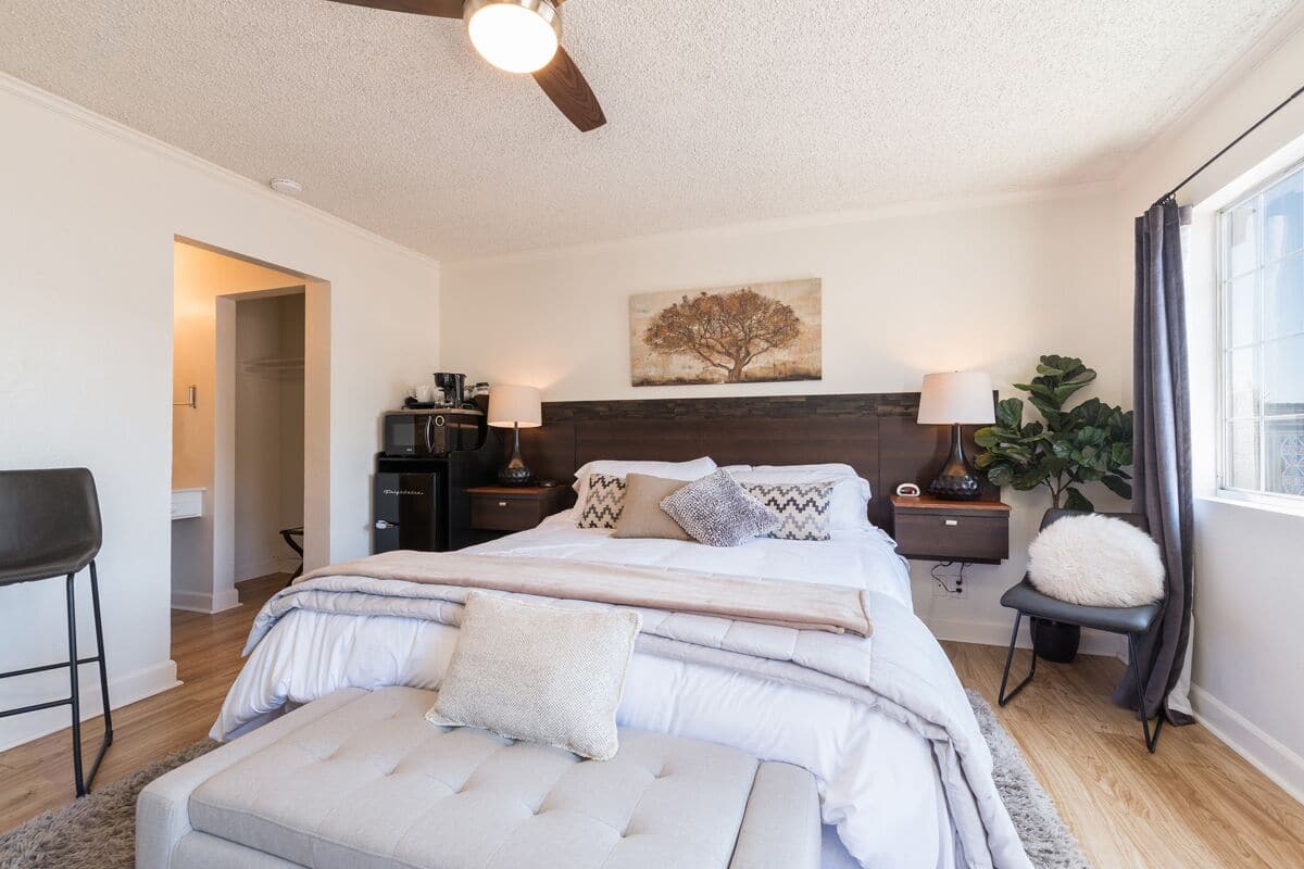 A bedroom with a large bed, a ceiling fan, a nightstand with a lamp, a wall-mounted shelf, and a window with curtains.