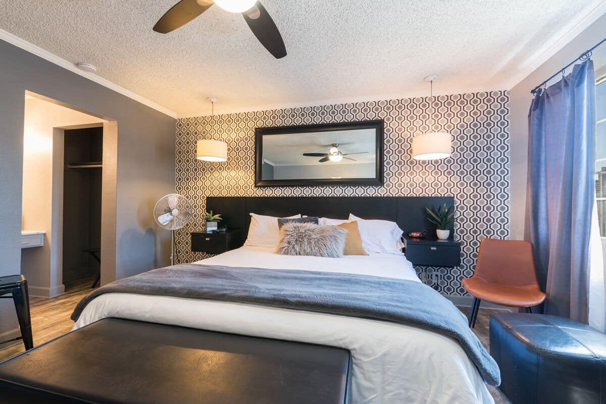A modern hotel room with a large bed, a ceiling fan, a mirror, and a small side table with a lamp.