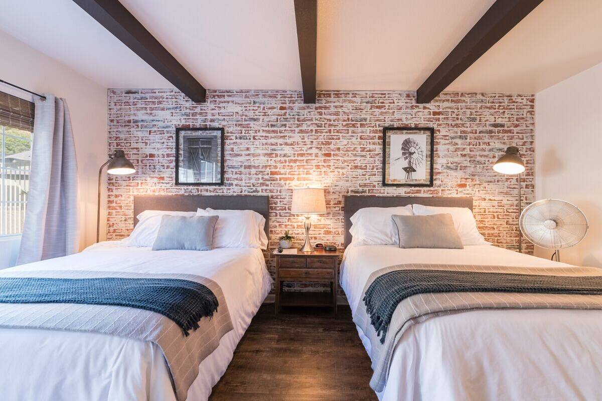 A hotel room with two beds, a brick wall, framed pictures, a nightstand with a lamp, and a fan.