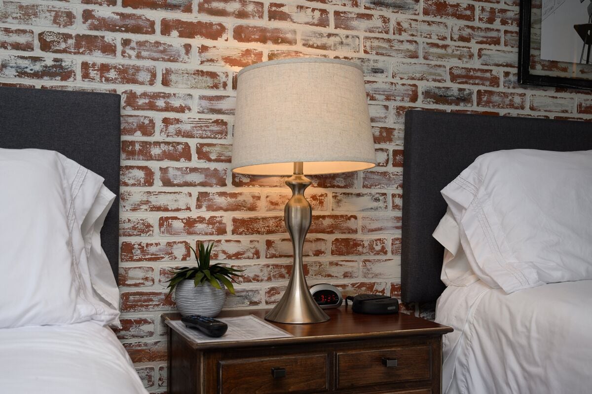 A hotel room with a brick-patterned wall, a bedside table with a lamp, a clock, and a potted plant.