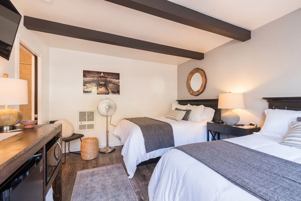 A hotel room with two beds, a desk with a lamp, a fan, and a wall-mounted mirror.