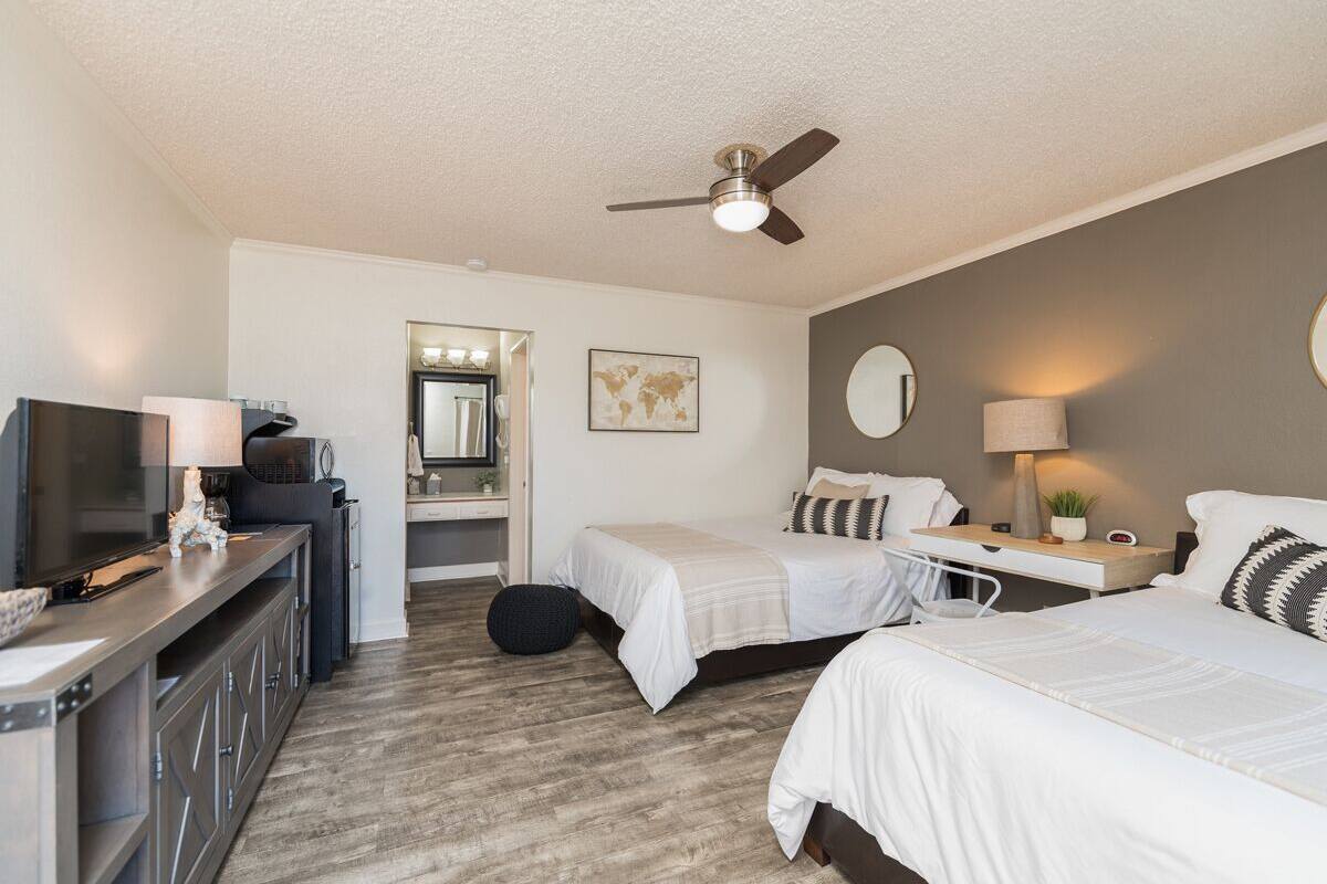 A hotel room with two beds, a desk, a TV, a ceiling fan, and a mirror.