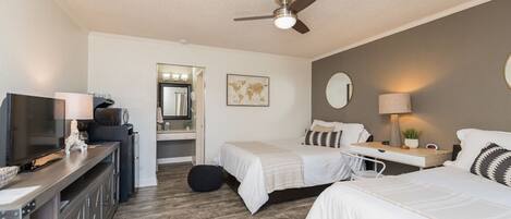 A hotel room with two beds, a desk, a TV, a ceiling fan, and a mirror.