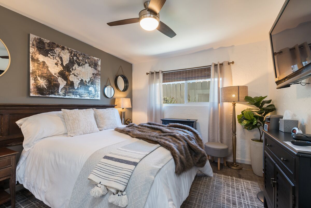 A bedroom with a bed, a ceiling fan, a world map artwork, a lamp, a window with curtains, and a bedside table.