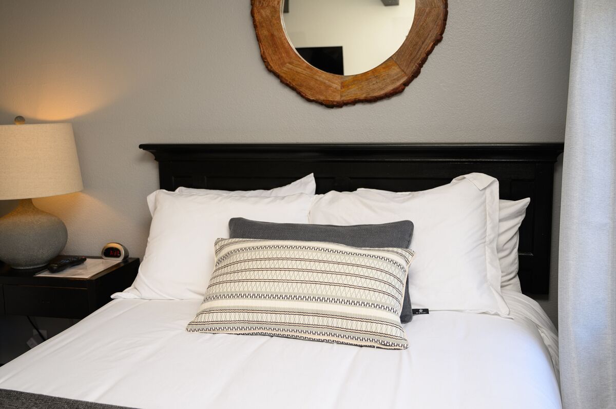 A neatly made bed with white bedding and a patterned pillow, a round mirror on the wall, a lamp on a bedside table, and a nightstand with a clock.