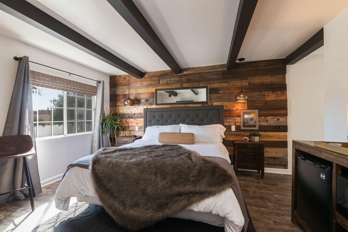 A bedroom with a large bed, wooden wall paneling, a small bedside table, a chair, and a window with curtains.