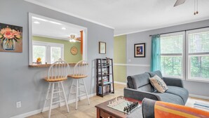 A living room with a grey sofa, a wooden coffee table, and a chessboard. There is a bookshelf with books and a mirror above a dining table with two chairs.