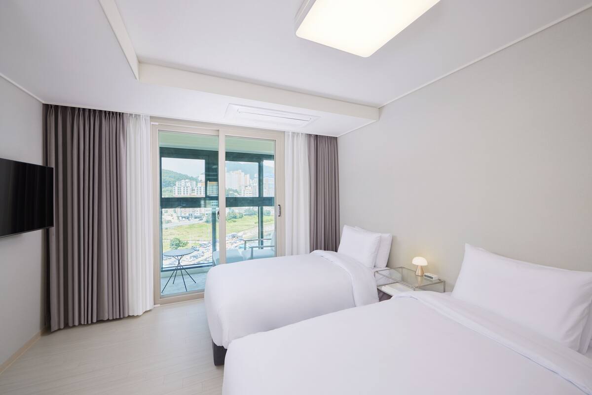 A hotel room with two beds, a TV mounted on the wall, a bedside table with a lamp, and a large window offering a city view.