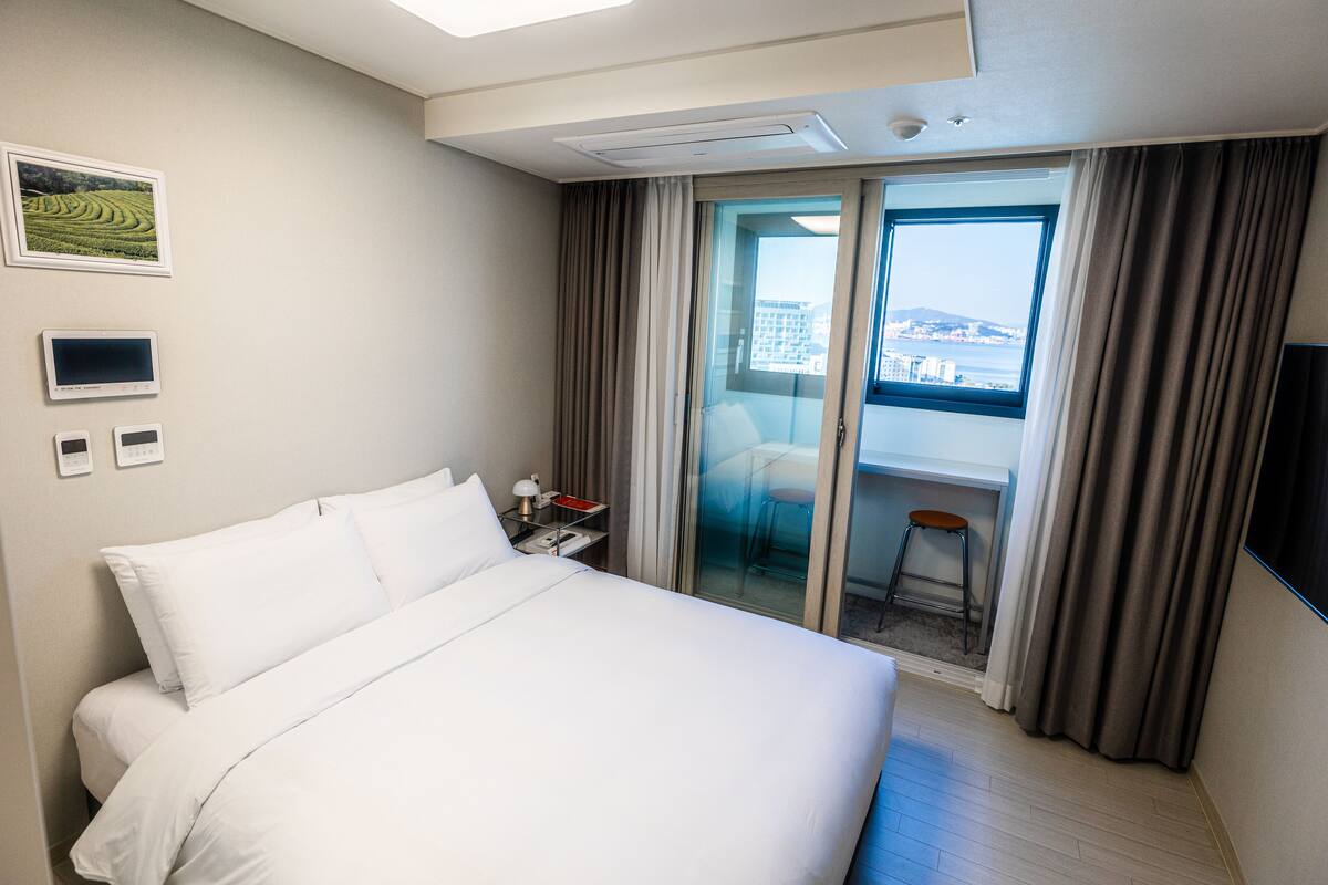 A hotel room with a large bed, a desk with a lamp, a TV, and a view of the city through a sliding door.