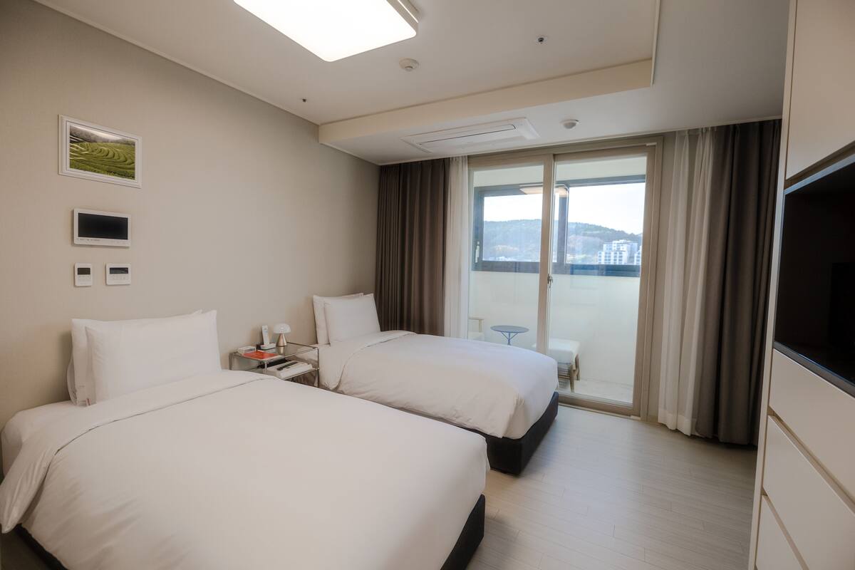 A hotel room with two beds, a TV, a bedside table, a window with curtains, and a view of buildings outside.