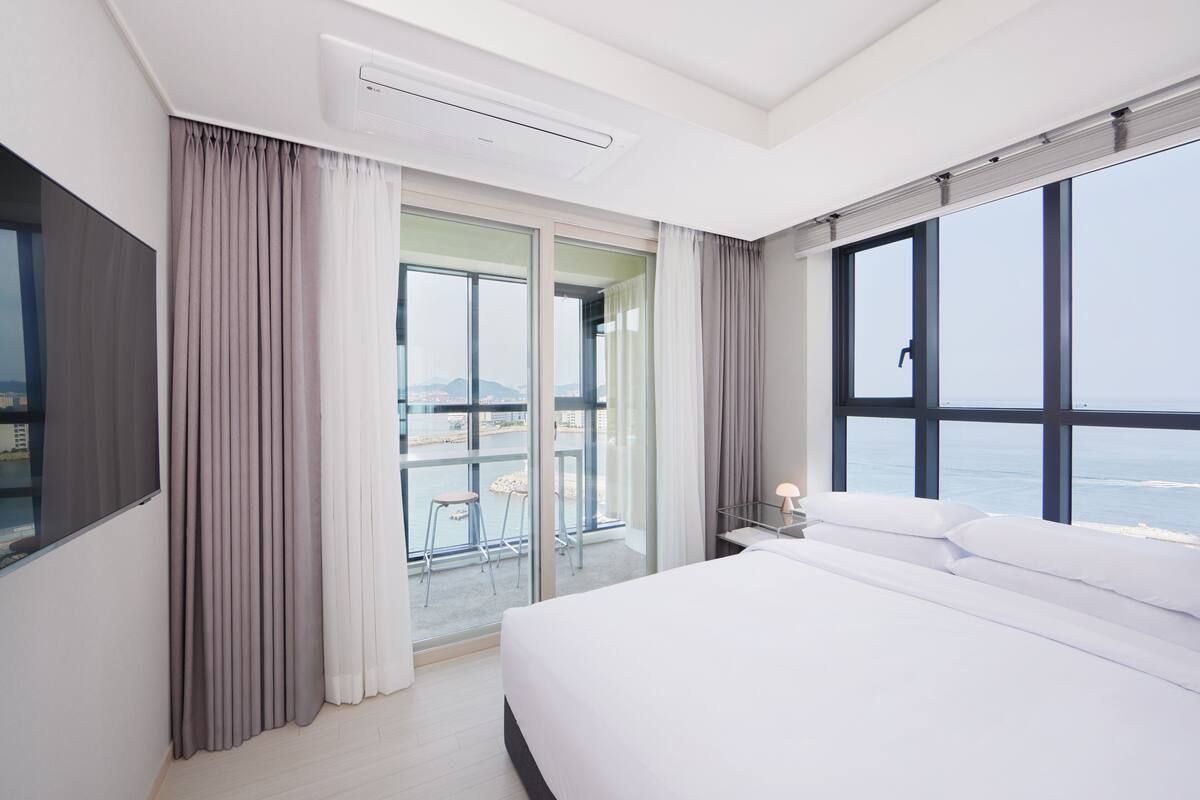 A modern hotel room with a large bed, a balcony with a view of the sea, and a sliding glass door.