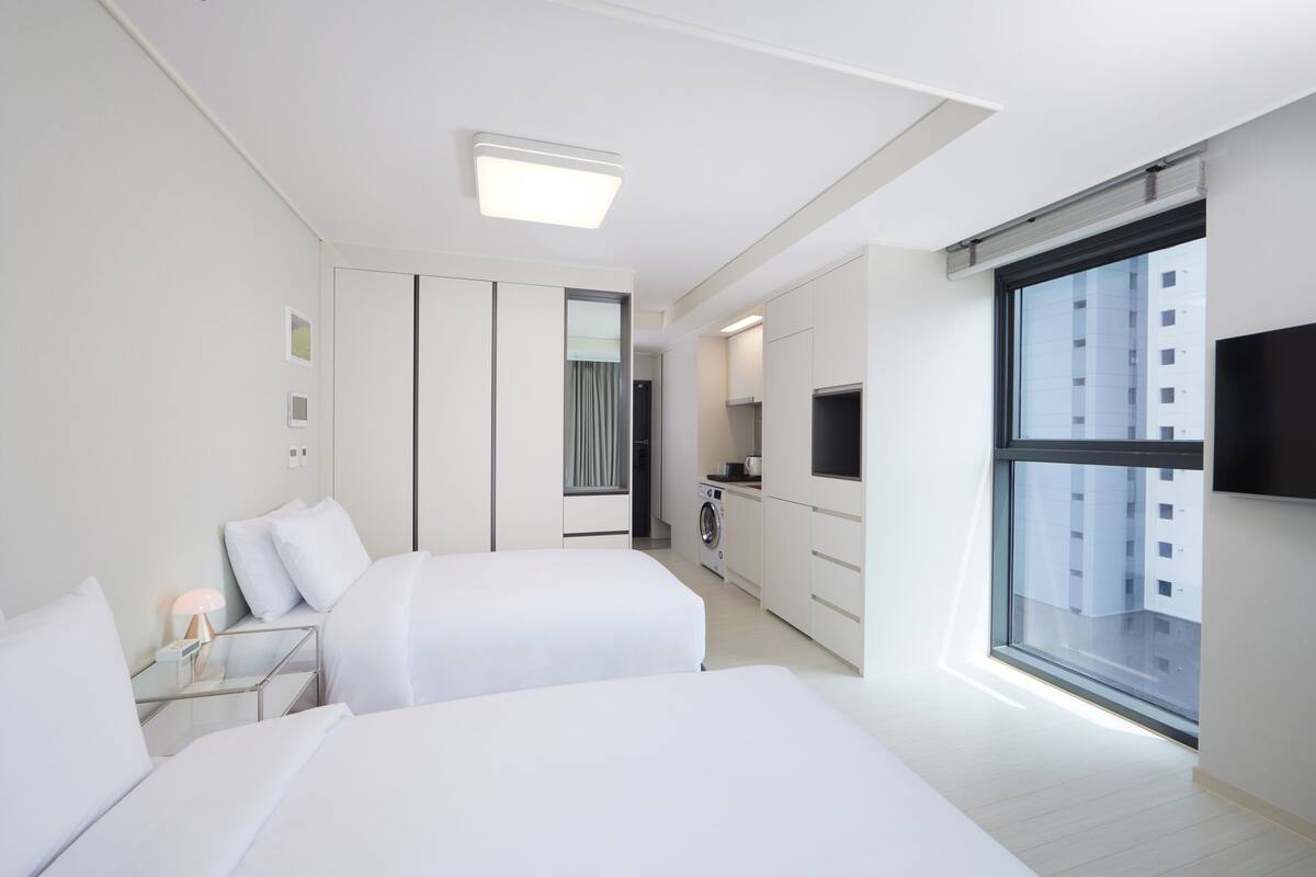 A modern hotel room with two beds, a small kitchenette, a washing machine, and a large window offering a city view.