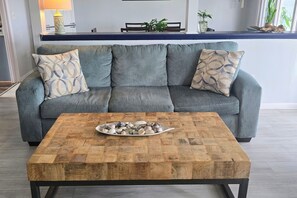 A grey sofa with patterned cushions, a wooden coffee table with a shell centerpiece, and a kitchen area in the background.