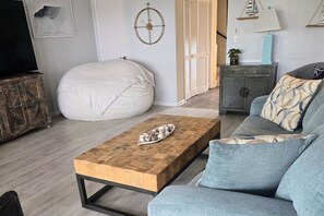 A living room with a wooden coffee table, a grey sofa, a round bean bag, a small cabinet, and a wall clock.