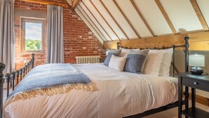 A bedroom with a sloped ceiling, exposed wooden beams, a brick wall, a large bed with a headboard, and a nightstand with a lamp. - The Cart Shed (NC) (4), North Creake, Norfolk (North Creake)
