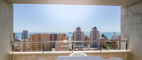 A balcony with a table and chairs, overlooking a cityscape with high-rise buildings and a clear sky.