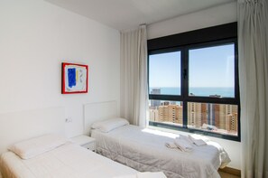 Een hotelkamer met een bed, een nachtkastje, een raam met uitzicht op gebouwen en de zee, en een schilderij aan de muur.