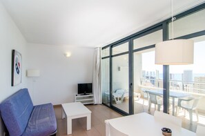 A modern hotel room with a balcony, a flat-screen TV, a sofa, a small table, and a view of the city.