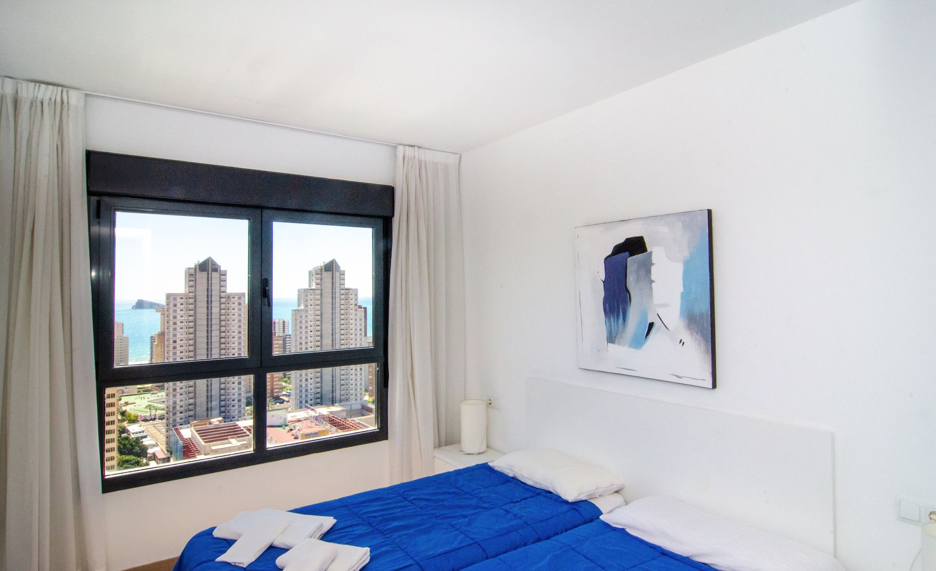 A hotel room with a bed, a window with curtains, a painting on the wall, and a view of buildings.