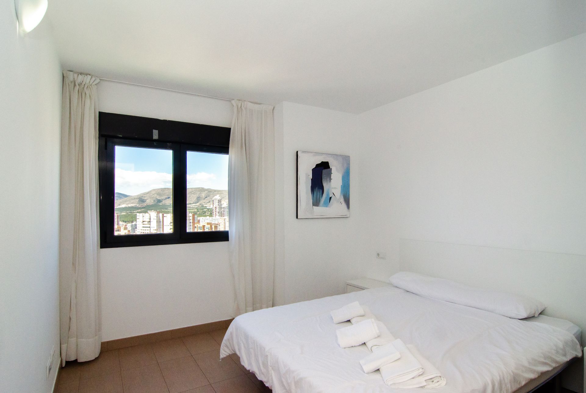 A hotel room with a bed, a window with curtains, a wall-mounted painting, and a view of buildings and hills.