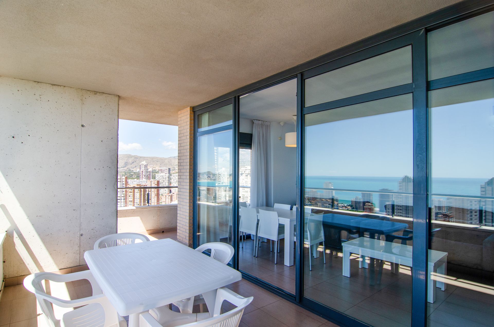 A balcony with a table and chairs, a glass door leading to an outdoor seating area, and a cityscape view.