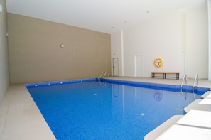 A large indoor swimming pool with clear blue water, a lifebuoy on the wall, and a bench near the pool.
