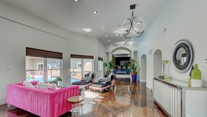 Un salón espacioso con un sofá rosa, una mesa de centro y una araña. Se ve una piscina y un comedor al fondo.