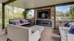 Un patio al aire libre con una chimenea de piedra, un televisor montado en la pared y zonas de estar cómodas.