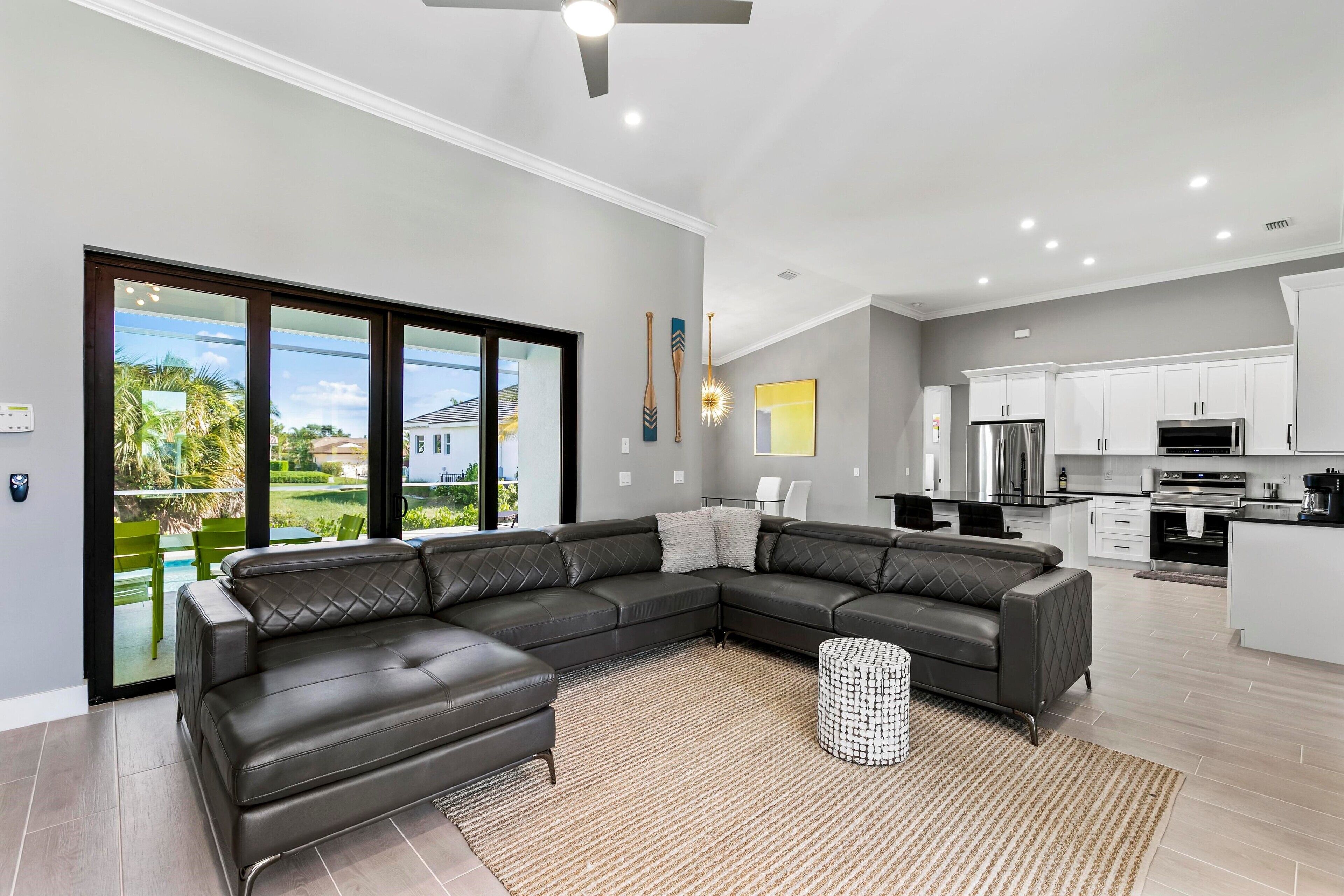 A modern living room with a sectional sofa, a kitchen with white cabinets, and a view of the outdoors.