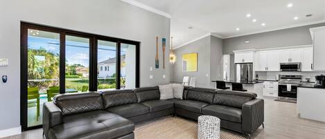 A modern living room with a sectional sofa, a kitchen with white cabinets, and a view of the outdoors.