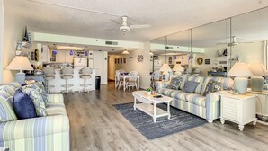 Un vaste salon avec un grand canapé, un coin repas avec des chaises, une cuisine à l’arrière-plan et un ventilateur de plafond.