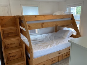 Una cama litera de madera con escalera, un pequeño escritorio y una ventana con persianas.