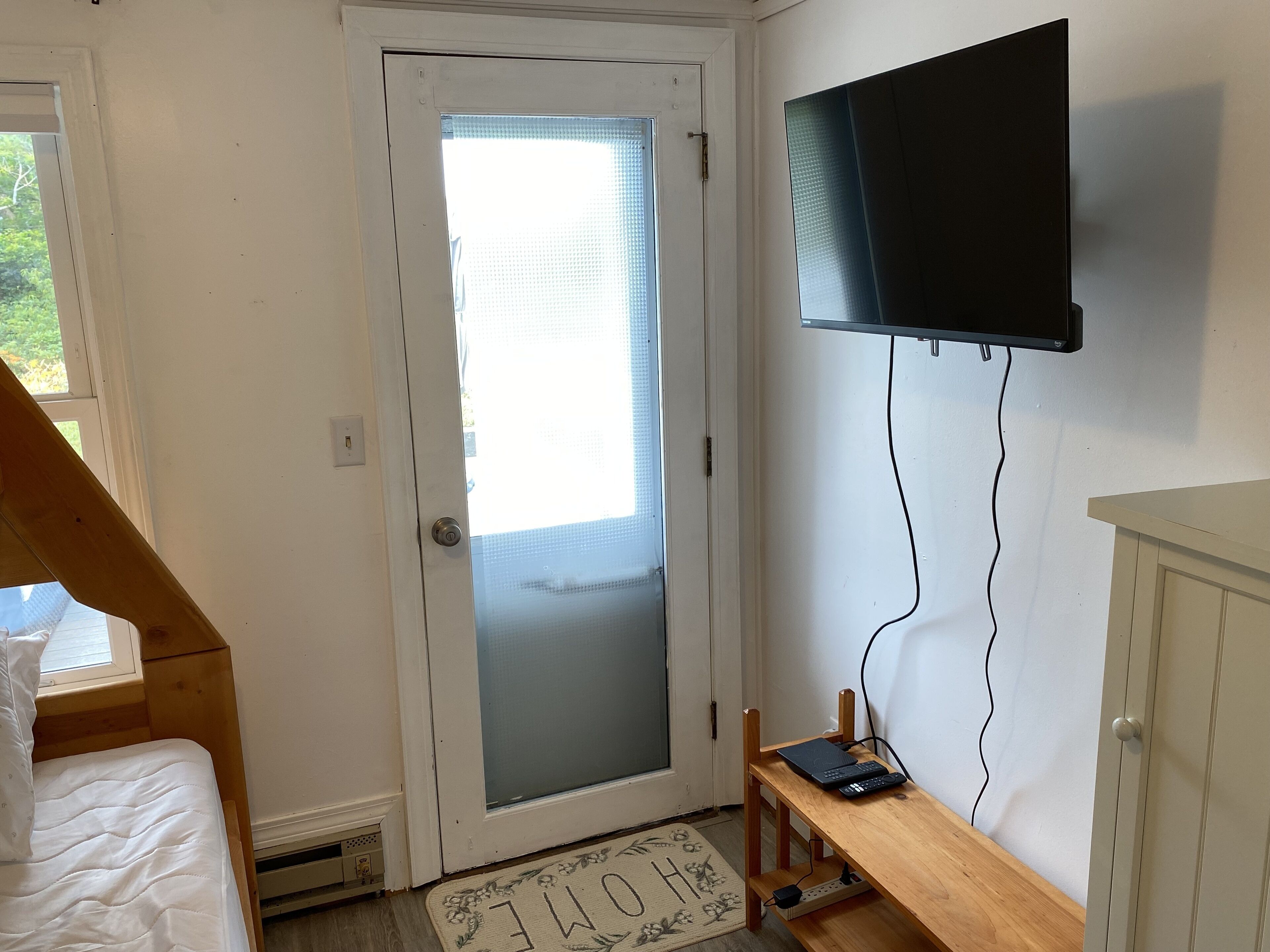 A bedroom with a bed, a desk with a laptop, a TV mounted on the wall, and a door with a frosted glass panel.