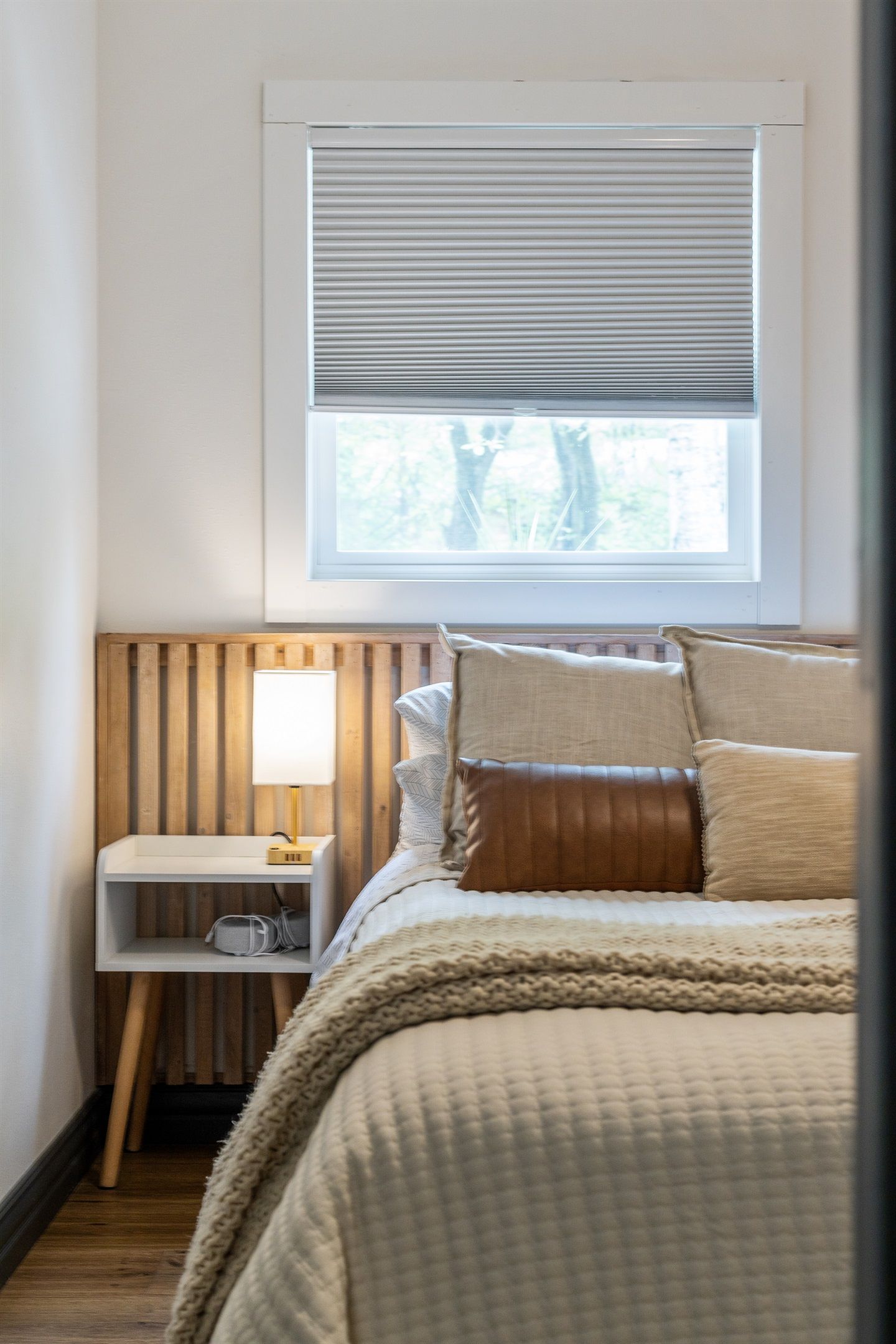 Un dormitorio con una cama, una mesita de noche con lámpara, una ventana con persianas y un cabecero de madera.