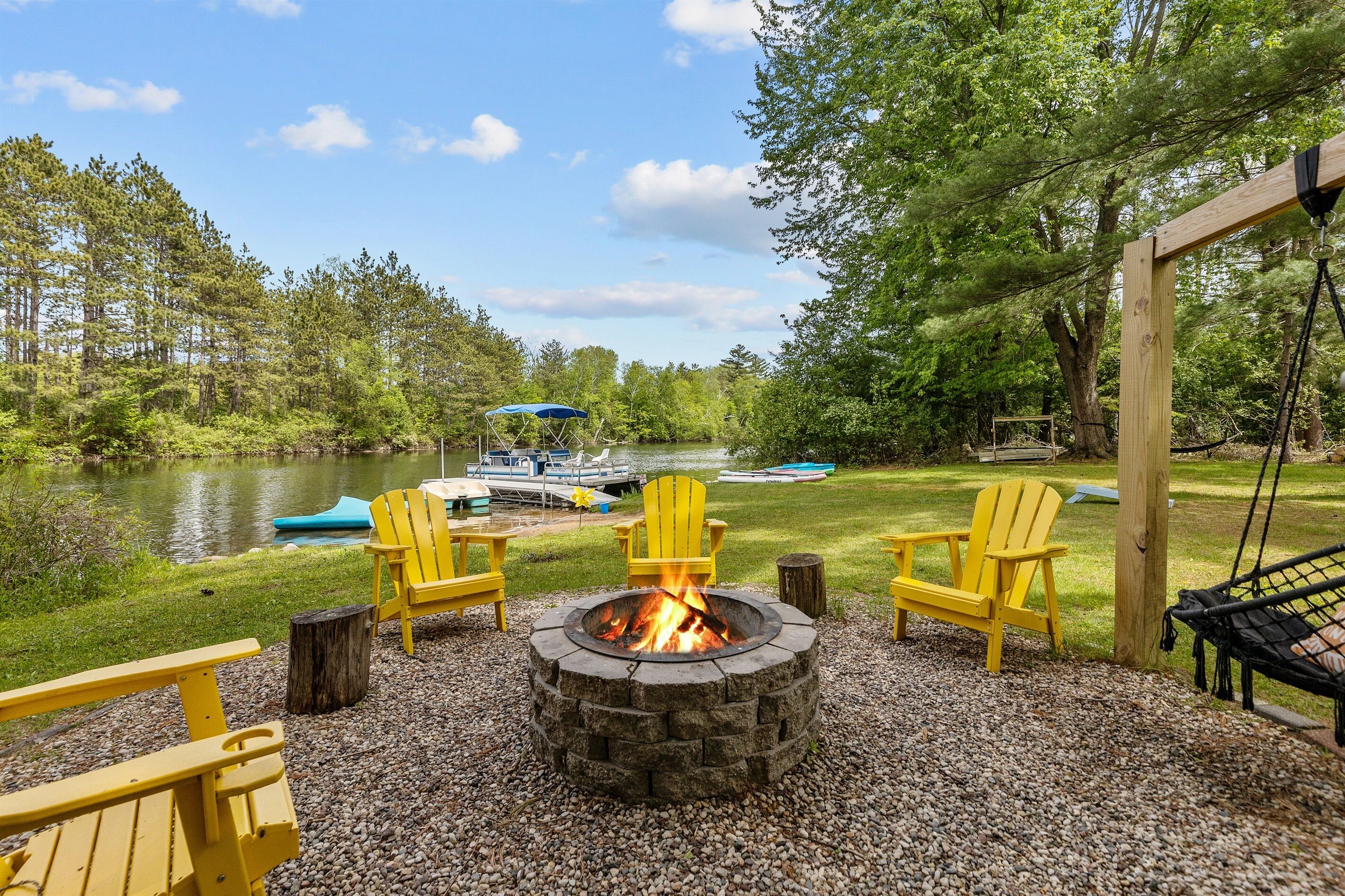 A fire pit surrounded by Adirondack chairs, a hammock, and a view of a lake with boats.