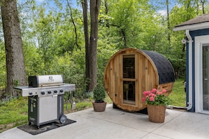 Uma sauna de madeira, uma churrasqueira e um pequeno galpão numa área arborizada.