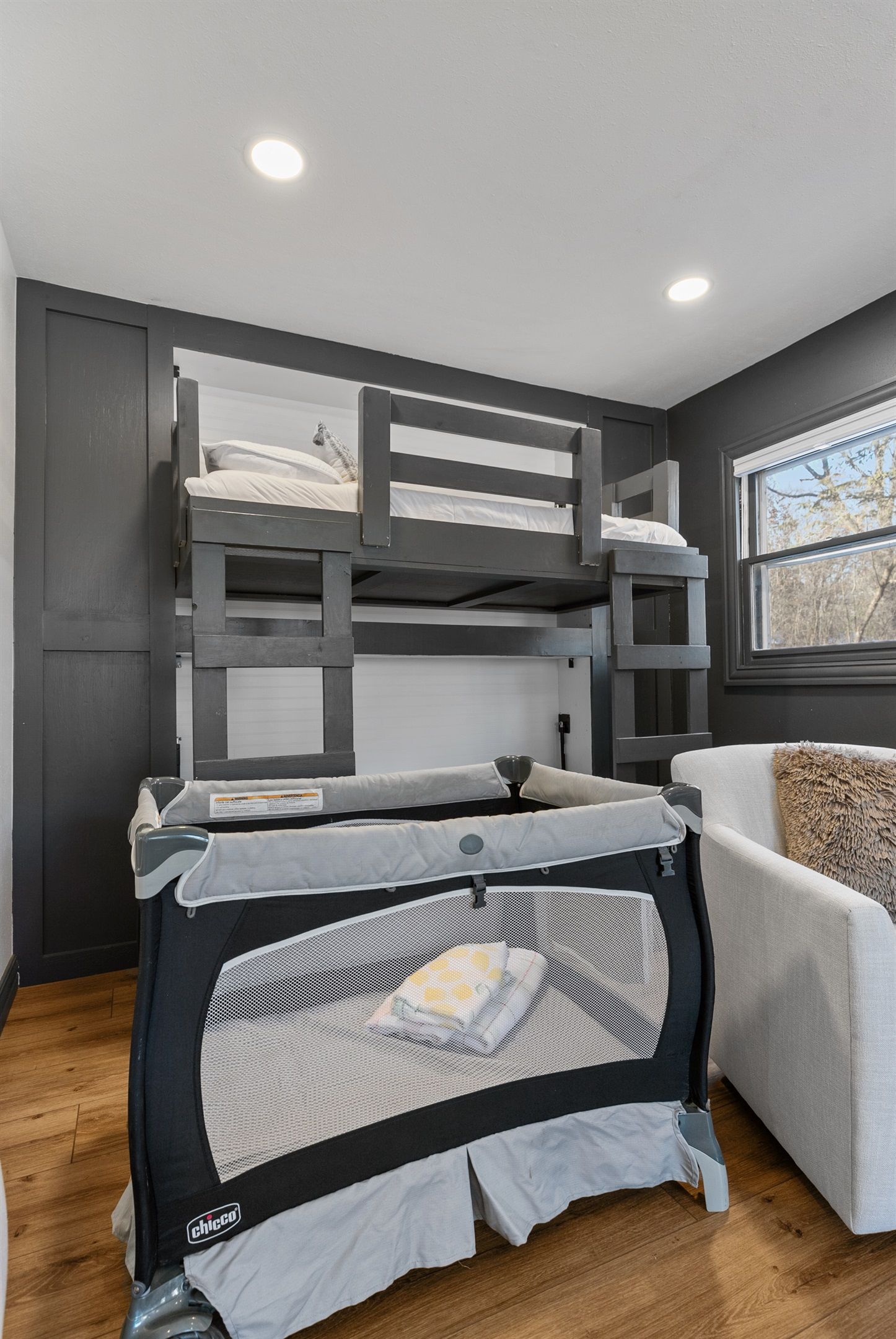 A bunk bed with a safety rail, a white couch, and a window with a view of trees.