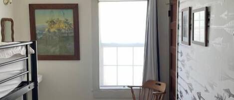 A room with a large window, a chair, a black metal rack, and a painting on the wall.
