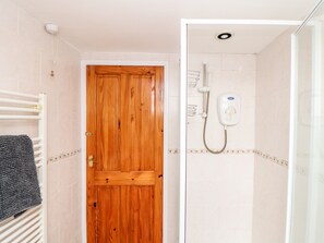 A bathroom with a wooden door, a white towel rack, a shower with a handheld showerhead, and a tiled floor. - Keepers Cottage (Liskeard)