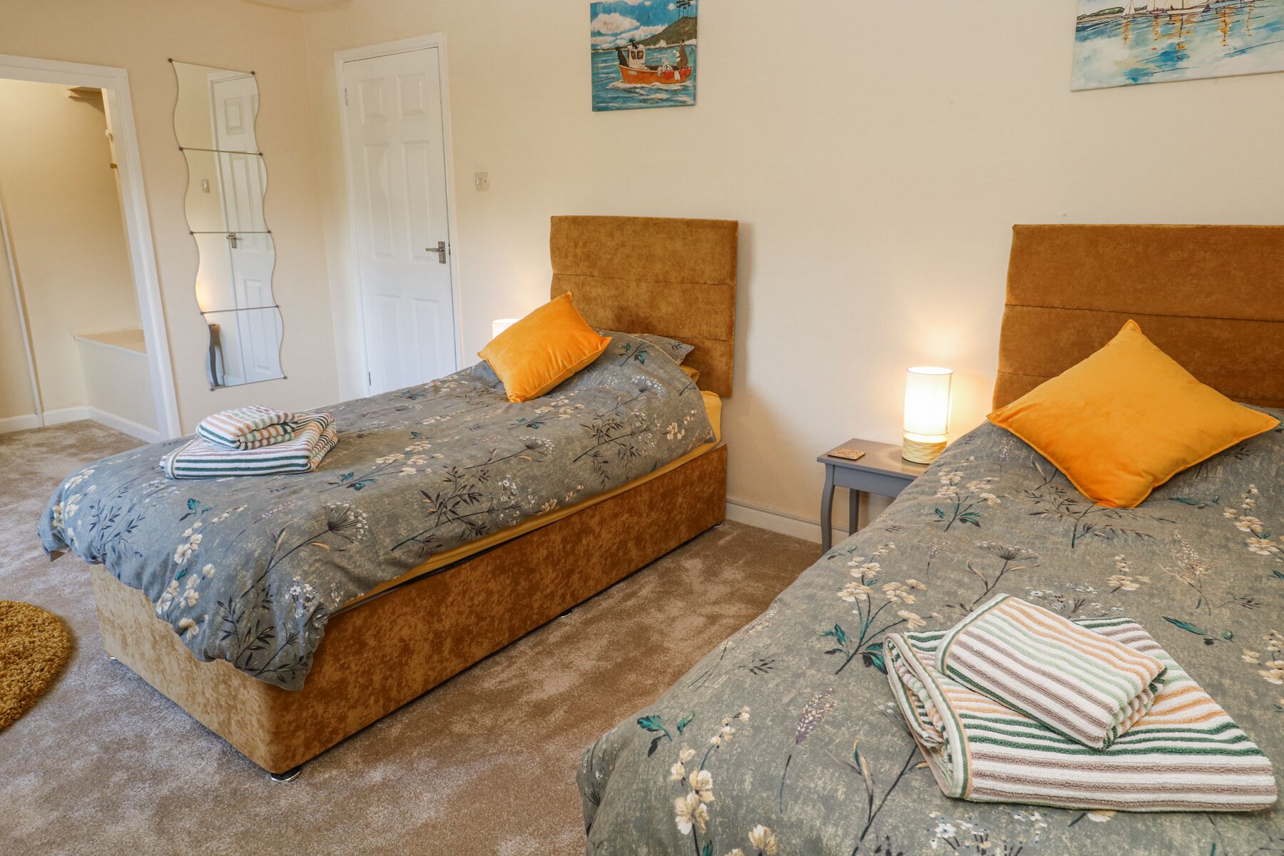 Un dormitorio con dos camas, cada una con ropa de cama floral y almohadas naranjas. Hay dos mesitas de noche con lámparas, una puerta y un espejo en la pared.