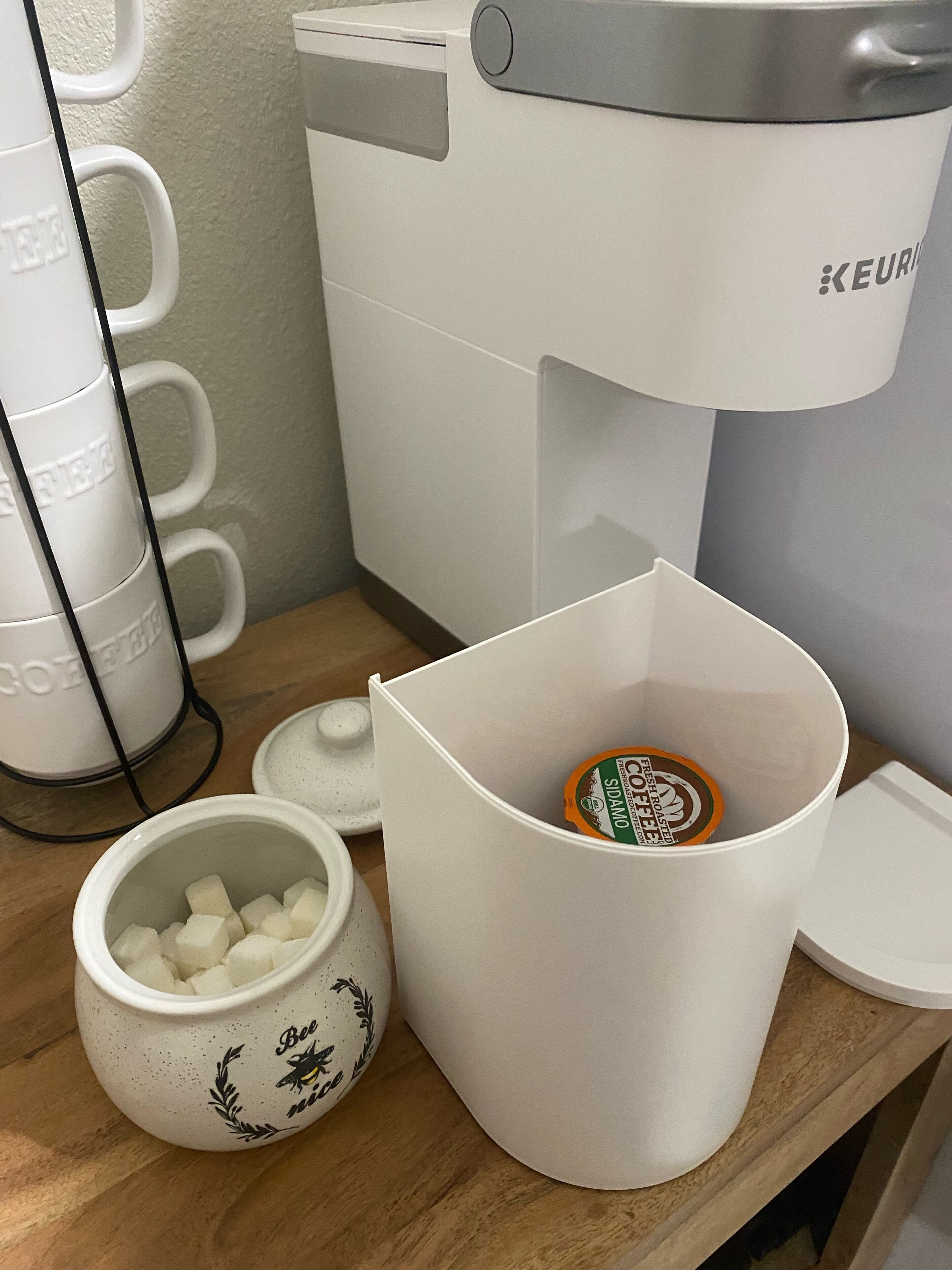 A coffee maker, a mug with sugar cubes, and a cup with a lid on a wooden surface.