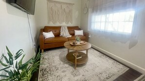 Un salon avec un canapé en cuir marron, une table basse ronde en bois, une décoration murale en macramé blanc et une grande fenêtre ornée de rideaux légers.