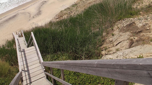 Een houten looppad naar een zandstrand met golven, begroeide duinen en een heldere lucht.